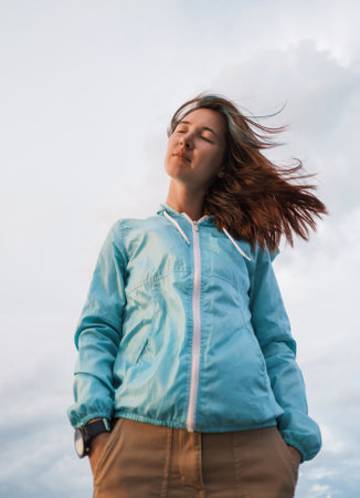 A pensive young woman stands in the cloudy sky background.の写真素材