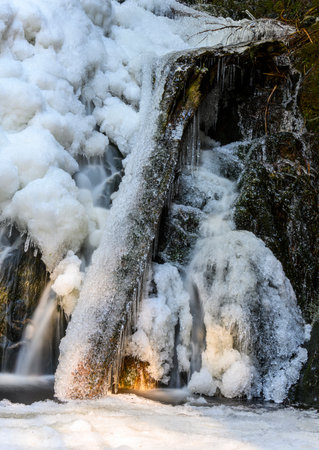 Beautiful frozen waterfall in the winter. Frozen water an ice everywhere. Icy wood log and rocks. Light rays shining through the woodsの写真素材