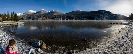 Stunning winter landscape panorama. Frozen lake, snow and snow capped mountains. Little girl in pink is in the foregroundの写真素材