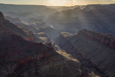 Sunrise illuminates the Grand Canyon, highlighting its rugged cliffs and valleys. The serene river...