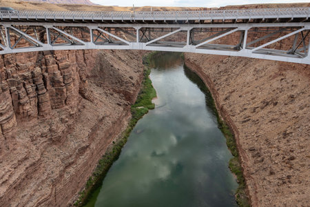 A bridge spans over a serene river winding through a dramatic canyon landscape. The reflection of clouds dances on the water's surface, creating a tranquil atmosphere.の写真素材