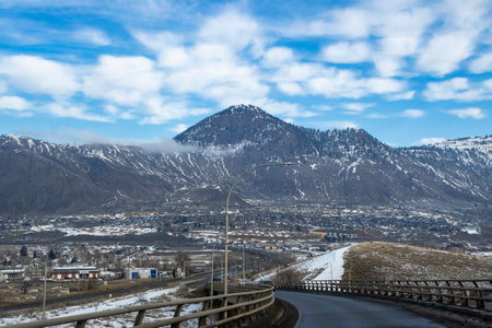 A winding road leads through a snow-covered area with a mountain backdrop. The sky features fluffy clouds, showcasing a serene, early spring day.の写真素材