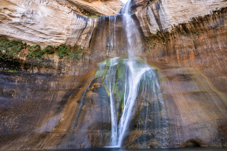 Water tumbles gracefully over rocky cliffs into a tranquil pool, surrounded by towering canyon walls and lush greenery under a bright midday sun.の写真素材