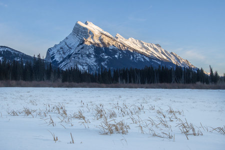 A majestic mountain peak adorned with snow stands tall against a clear twilight sky. In the foreground, a frozen landscape with patches of grass contrasts the towering nature.の写真素材