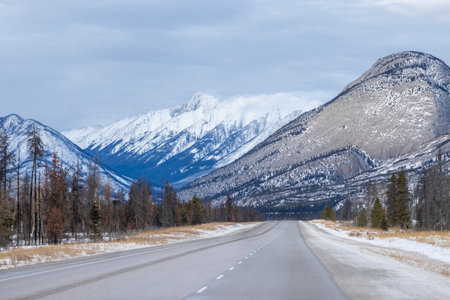A scenic view showcases snow-capped mountains rising majestically in the distance. A smooth, winding road leads through a tranquil winter landscape under moody clouds.の写真素材