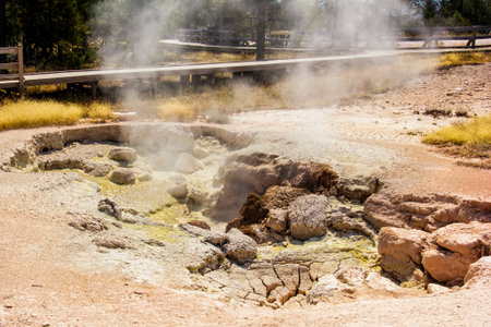 A colorful geothermal feature releases steam, surrounded by earthy textures and a wooden pathway nearby. Visitors can admire the unique landscape and vibrant colors of the natural setting.の写真素材