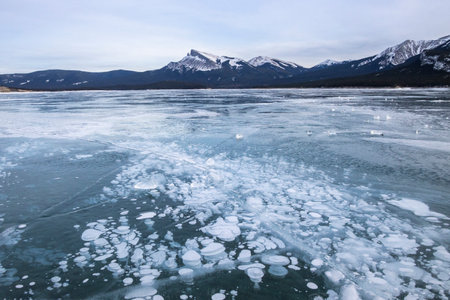 Crystal clear ice bubbles create an intriguing pattern on a frozen lake, surrounded by snow-capped mountains under a calm winter sky. The serene atmosphere invites exploration.の写真素材