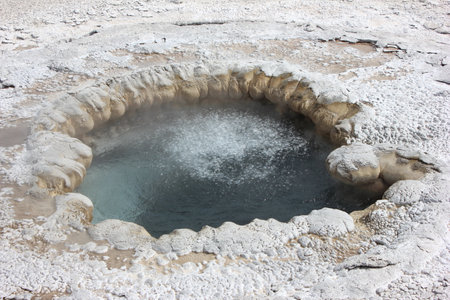 Bubbling geyser creates steam in Yellowstone National Park under bright daylight. Mineral-rich deposits outline the thermal feature, showcasing nature's unique beauty.の写真素材