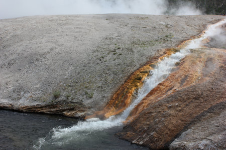 A bright orange hot spring cascades down rocky terrain into a serene body of water, surrounded by steam and a rugged landscape in Yellowstone National Park under grey skies.の写真素材