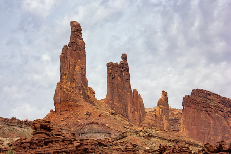 The striking rock formations tower above the desert terrain, illuminated by soft, diffused light under an overcast sky. Desert vegetation and rugged cliffs surround the area.の写真素材