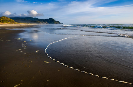 Soft waves wash ashore, creating intricate patterns on the sandy beach. The sun sets behind distant hills, painting the sky in warm hues while inviting calm and reflection.の写真素材