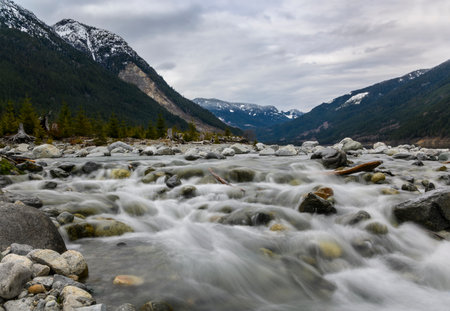 A river cascades smoothly over rocks, framed by towering mountains topped with snow under a cloudy sky. The landscape evokes a peaceful, natural atmosphere perfect for outdoor exploration.の写真素材