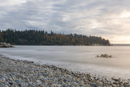 Gentle waves lap against a rocky shoreline as the sun sets, casting a soft glow on the pebbles. A...