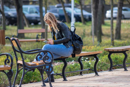 TIMISOARA, ROMANIA - NOVEMBER 17, 2019: Woman sitting on a bench and checking her phone. Real people.のeditorial素材