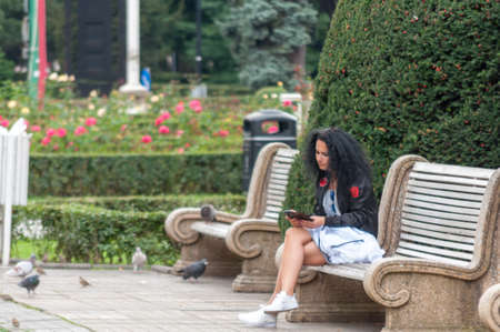 TIMISOARA, ROMANIA - SEPTEMBER 27, 2016: Woman sitting on a bench in the street and checking her phone. Real people.のeditorial素材