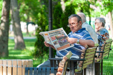 Timisoara, Romania - Jne 02, 2017: Man sitting on a bench and reading a newpaper in a park. Real people.のeditorial素材