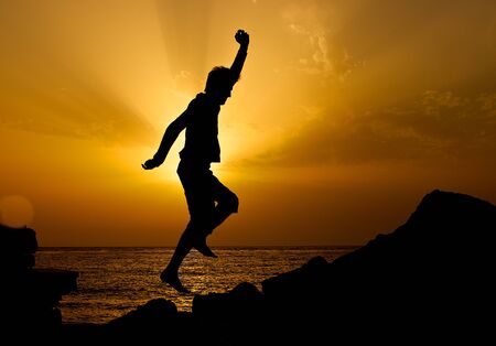 Silhouette of the jumping man on sunset sun. Jumping man near the sea. Happy man. Silhouette of man jumping on sunset. Unknown man on sunset. Sunset. Man on holidaysの写真素材