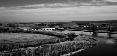 Monochrome of a prairie with a river crossed by a bridge in the distance.