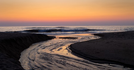 A water stream ends in the ocean with the sunset colors being reflected in the water.の写真素材