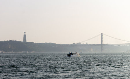 A watercraft sails in the Tagus River with the 25 de Abril Bridge and Cristo Rei in the background.