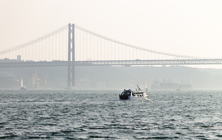 A watercraft sails in the Tagus River with the 25 de Abril Bridge and Cristo Rei in the background.の写真素材