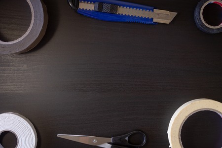 A black wooden background with various cutting tools: scissors, utility knife, tape.
