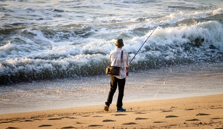 Fisherman stands with a fishing pole near the incoming ocean waves at the beach. Fisherman's hat, side bag. Rear view. Warm light.の写真素材