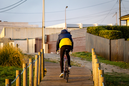 A female cyclist rides her bike on a boardwalk. Yellow and blue jacket. Back View. Cloudy day, warm light.の写真素材