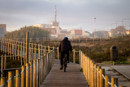 A cyclist rides his bike on a boardwalk near vegetation. Black jacket. Back View. Cloudy day, warm light.の写真素材