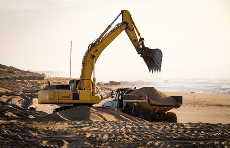 Yellow excavator raises its bucket while dump truck holds sand at the beach. Warm light background.