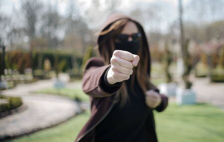 Young girl giving punch on unfocused background. Aggressive girl in a medical mask and glasses attacks.の写真素材