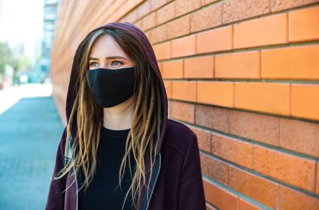 Girl with dreadlocks in a black protective mask on a background of an orange brick wall. coronavirus quarantine time. Copy space.の写真素材
