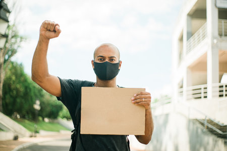 Dark skinned young man in a black mask holds a blank poster in one hand, while the other hand is...