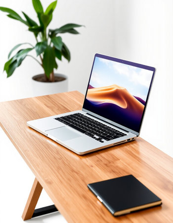 Laptop on a wooden desk with a notebook and a plant in the background