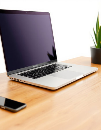 Laptop on wooden desk with smartphone and plant in the background.