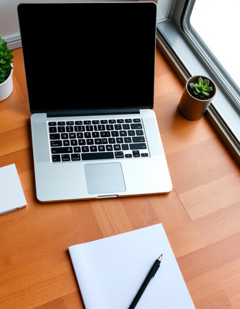 Laptop with blank screen and notebook on wooden table near window.