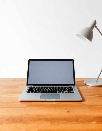 Laptop with blank screen on wooden table against white wall with lamp
