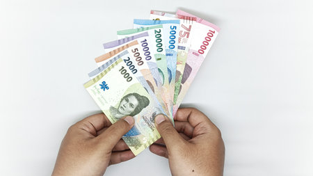 two hands holding a collection of Indonesian banknotes in denominations 1000, 2000, 5000, 10000,...