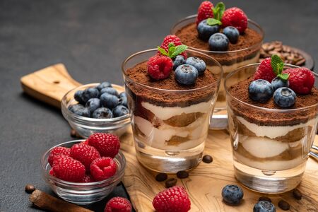 Classic tiramisu dessert with blueberries and raspberries in a glass on wooden cutting board on dark concrete backgroundの写真素材