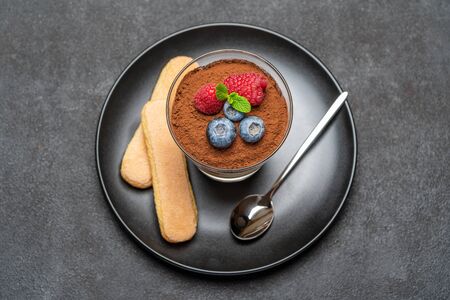 Classic tiramisu dessert with blueberries and raspberries in a glass and savoiardi cookies on dark concrete backgroundの写真素材
