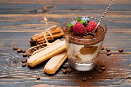 Classic tiramisu dessert portion with blueberries and strawberries in a glass cup and savoiardi cookies on wooden backgroundの写真素材