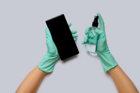 Females hands in latex gloves wiping mobile phone with alcohol decontaminating agent top view over light grey backgroundの写真素材
