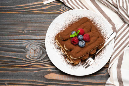 portion of Classic tiramisu dessert with raspberries and blueberries on ceramic plate isolated on wooden backgroundの写真素材