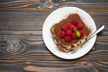 portion of Classic tiramisu dessert with raspberries on ceramic plate on wooden background