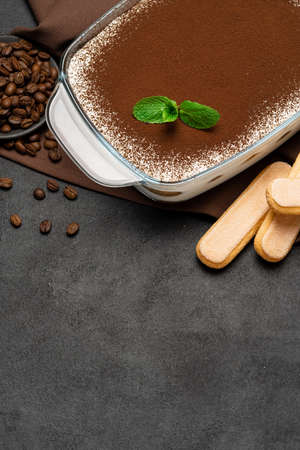 Traditional Italian Tiramisu dessert in glass baking dish and savoiardi cookies on concrete...