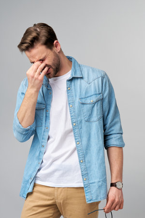 Frustrated young caucasian man in jeans shirt touching head with hand while standing overg studio grey backgroundの写真素材
