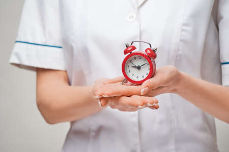 smiley nurse or doctor pointing holding small red alarm clock satanding over grey backgroundの写真素材