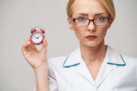 smiley nurse or doctor pointing holding small red alarm clock satanding over grey backgroundの写真素材