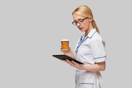 Portrait of a female doctor holding paper cup of coffee and holding tablet pc pad standing over grey...