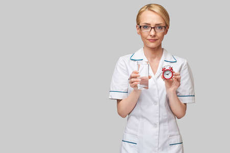 Healthy eating or lifestyle concept - female woman doctor holding and a glass of clear fresh waterand small red alarm clockの写真素材